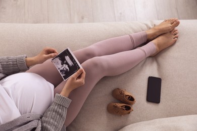 Young pregnant woman with ultrasound picture of baby on sofa, above view