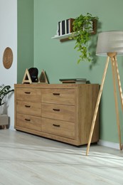 Photo of New wooden chest of drawers, plants and lamp near green wall in stylish room