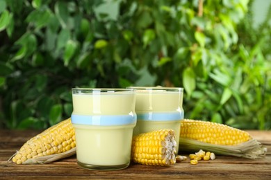 Photo of Tasty fresh corn juice in glasses and cobs on wooden table against blurred background