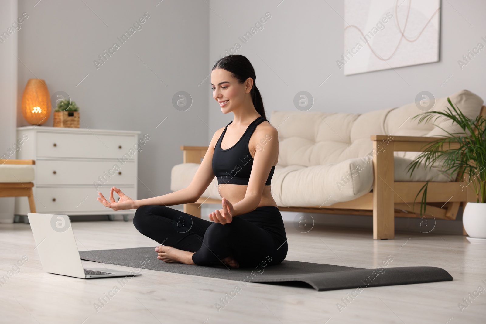Photo of Beautiful young woman practicing Padmasana while watching online class on yoga mat at home. Lotus pose