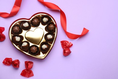 Heart shaped box with delicious chocolate candies and ribbon on lilac background, flat lay. Space for text