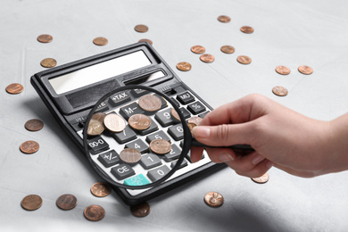 Woman looking through magnifying glass at calculator with coins on light grey table, closeup. Search concept