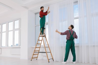 Photo of Workers in uniform hanging window curtain and showing thumbs up indoors