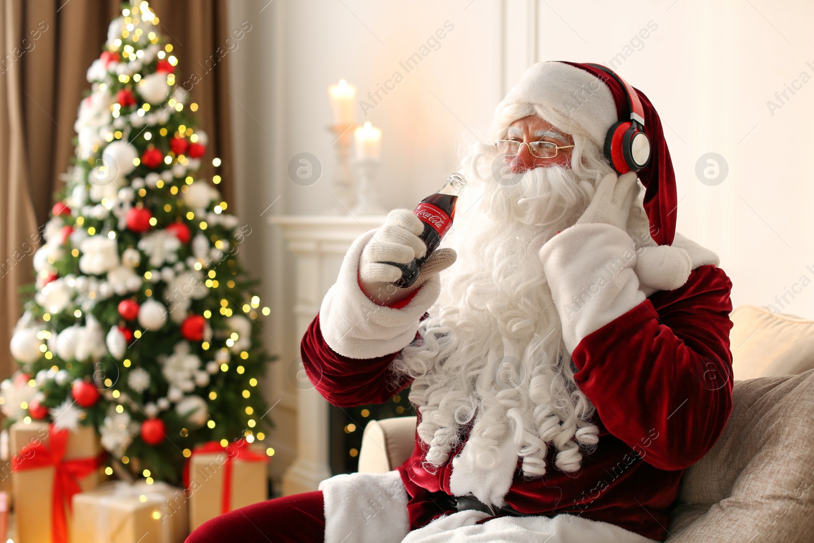 Photo of MYKOLAIV, UKRAINE - JANUARY 18, 2021: Santa Claus holding Coca-Cola bottle and listening to music with headphones in room decorated for Christmas