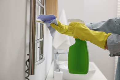 Photo of Woman cleaning heated towel rail with sprayer and rag, closeup
