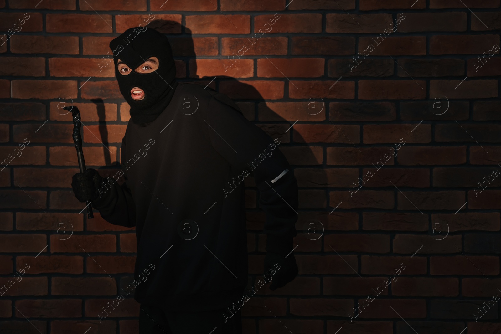 Photo of Thief in balaclava with crowbar against red brick wall