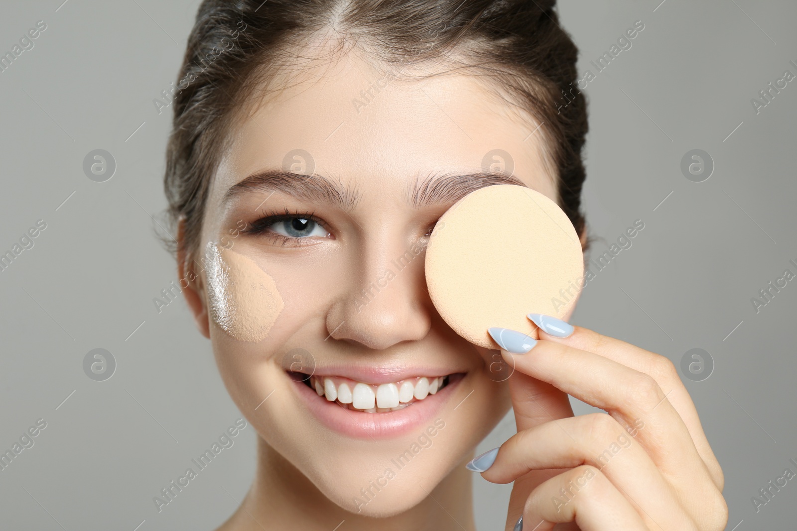 Photo of Beautiful girl with foundation smear on her face holding sponge against grey background, closeup