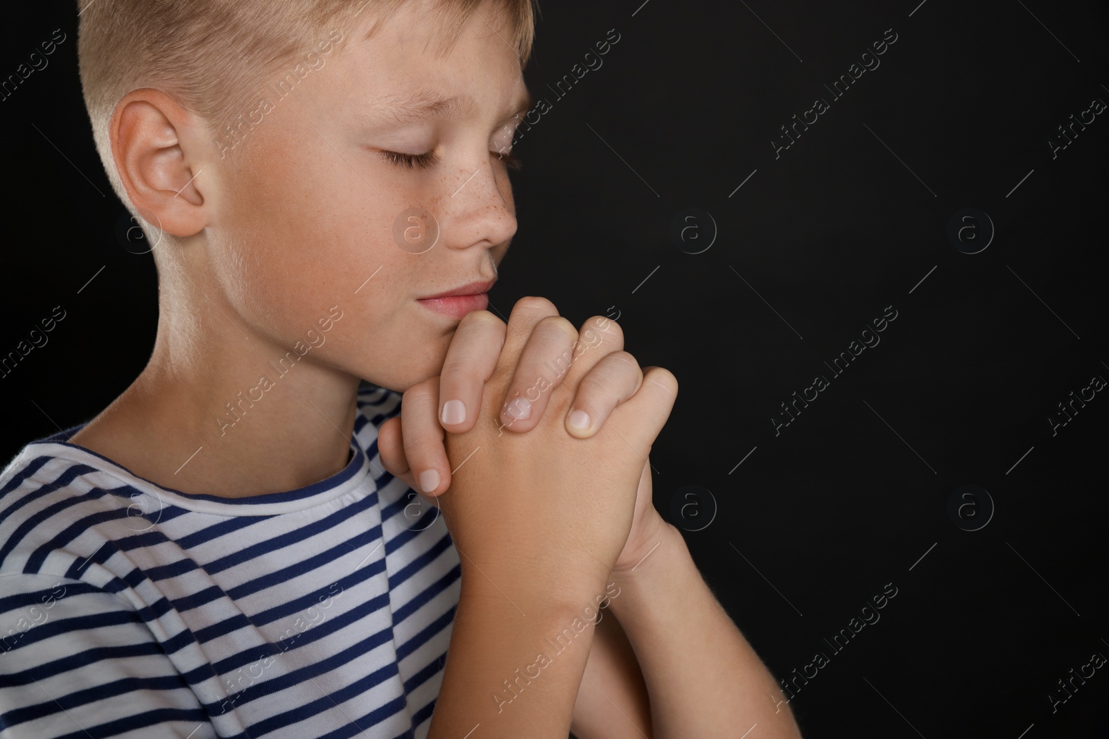 Photo of Boy with clasped hands praying on black background, closeup. Space for text