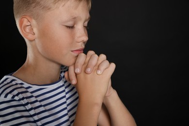 Photo of Boy with clasped hands praying on black background, closeup. Space for text