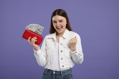 Excited woman holding wallet with dollar banknotes on purple background