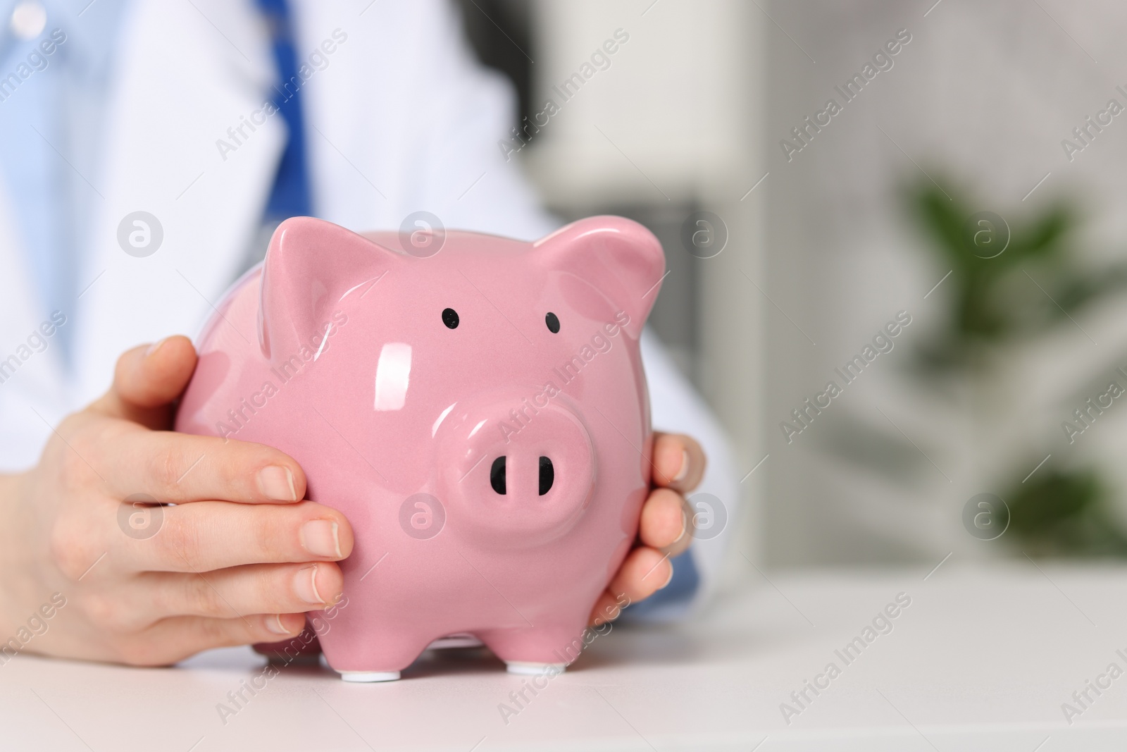 Photo of Doctor with piggy bank at white table indoors, closeup. Space for text