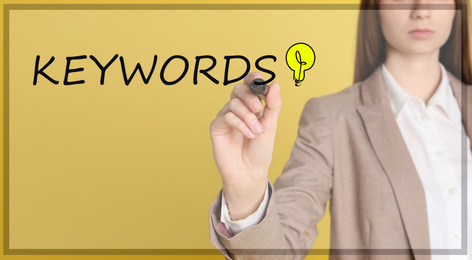Woman writing word KEYWORDS on transparent board against yellow background, closeup