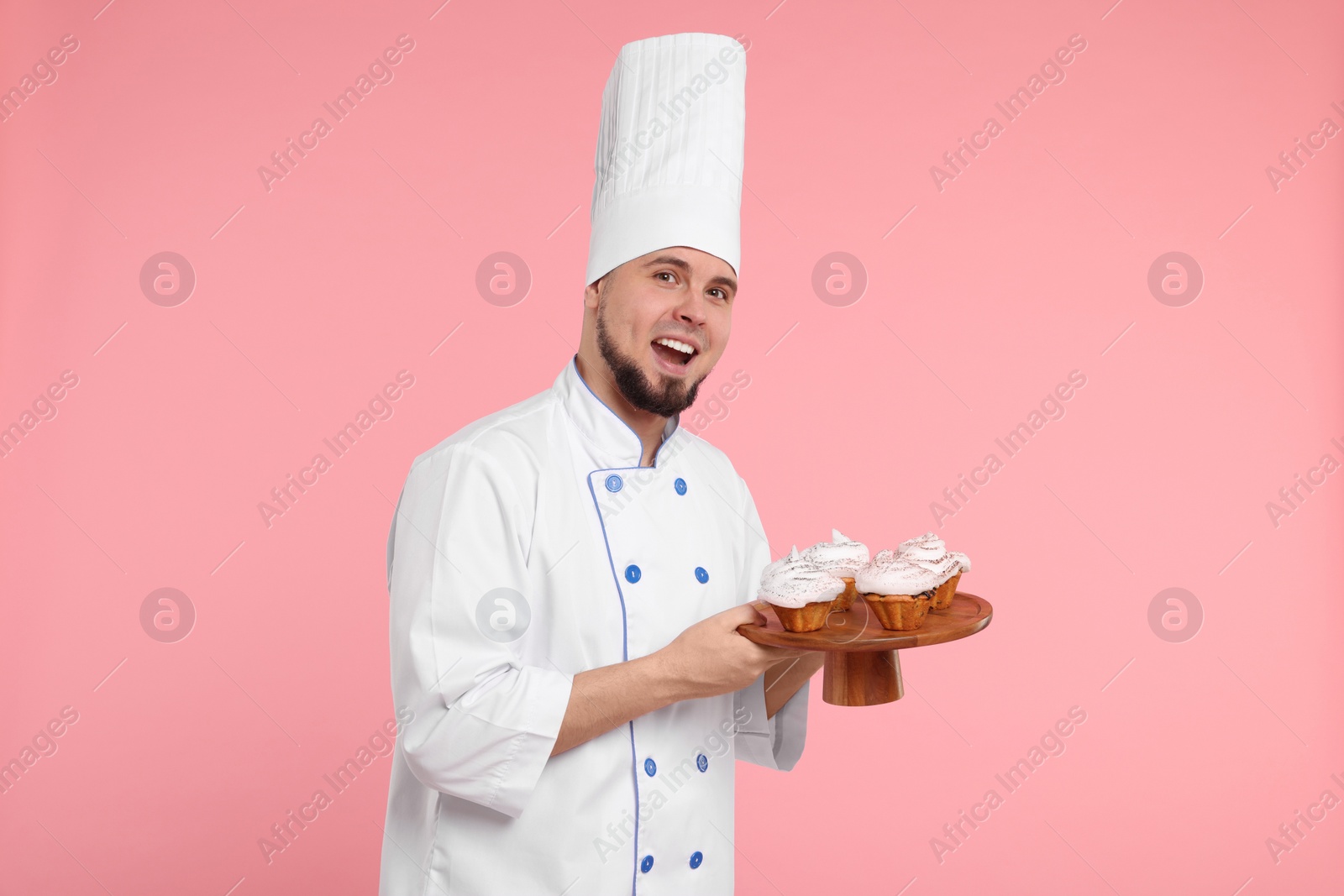 Photo of Happy professional confectioner in uniform holding delicious cupcakes on pink background