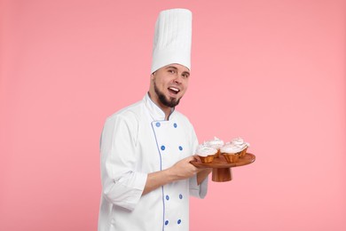 Photo of Happy professional confectioner in uniform holding delicious cupcakes on pink background