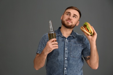 Young man with beer eating tasty burger on grey background. Space for text