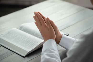 Woman holding hands clasped while praying at grey wooden table with Bible, closeup