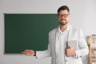 Happy teacher with book explaining something at blackboard in classroom