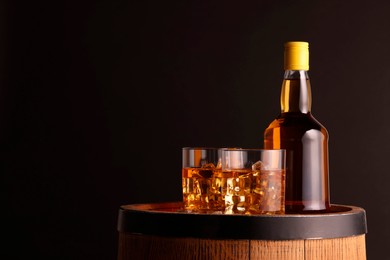 Whiskey with ice cubes in glasses and bottle on wooden barrel against black background, space for text