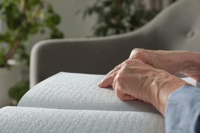 Photo of Blind senior person reading book written in Braille on sofa indoors, closeup