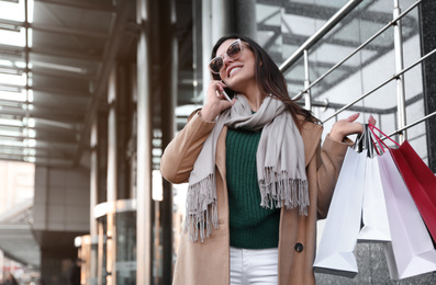 Photo of Beautiful young woman with shopping bags talking on smartphone outdoors