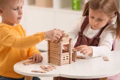 Photo of Little boy and girl playing with wooden house at white table indoors. Children's toys