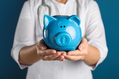 Photo of Doctor holding piggy bank on blue background, closeup. Medical insurance
