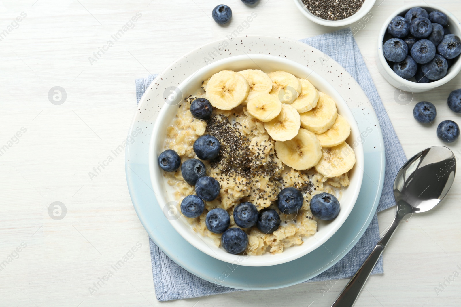 Photo of Tasty oatmeal with banana, blueberries and chia seeds served in bowl on white wooden table, flat lay