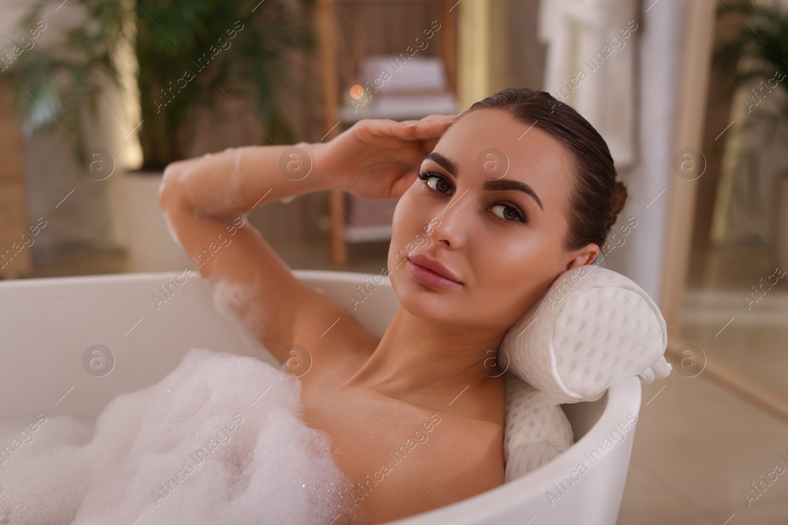 Photo of Young woman using pillow while enjoying bubble bath indoors