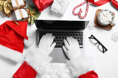 Photo of Santa Claus using laptop, closeup. Gift boxes, eyeglasses and Christmas decor on white table, top view