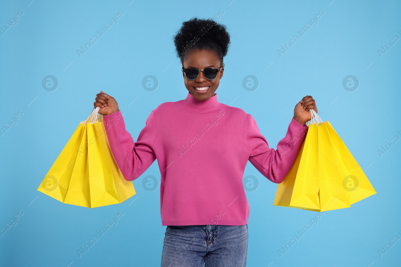 Photo of Happy young woman in stylish sunglasses with shopping bags on light blue background