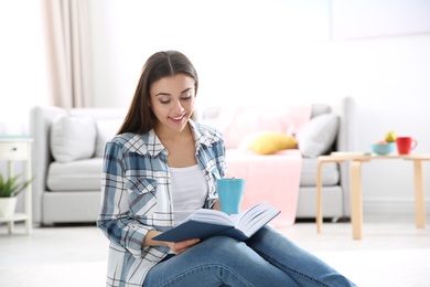 Photo of Beautiful young woman with cup of coffee reading book on floor at home