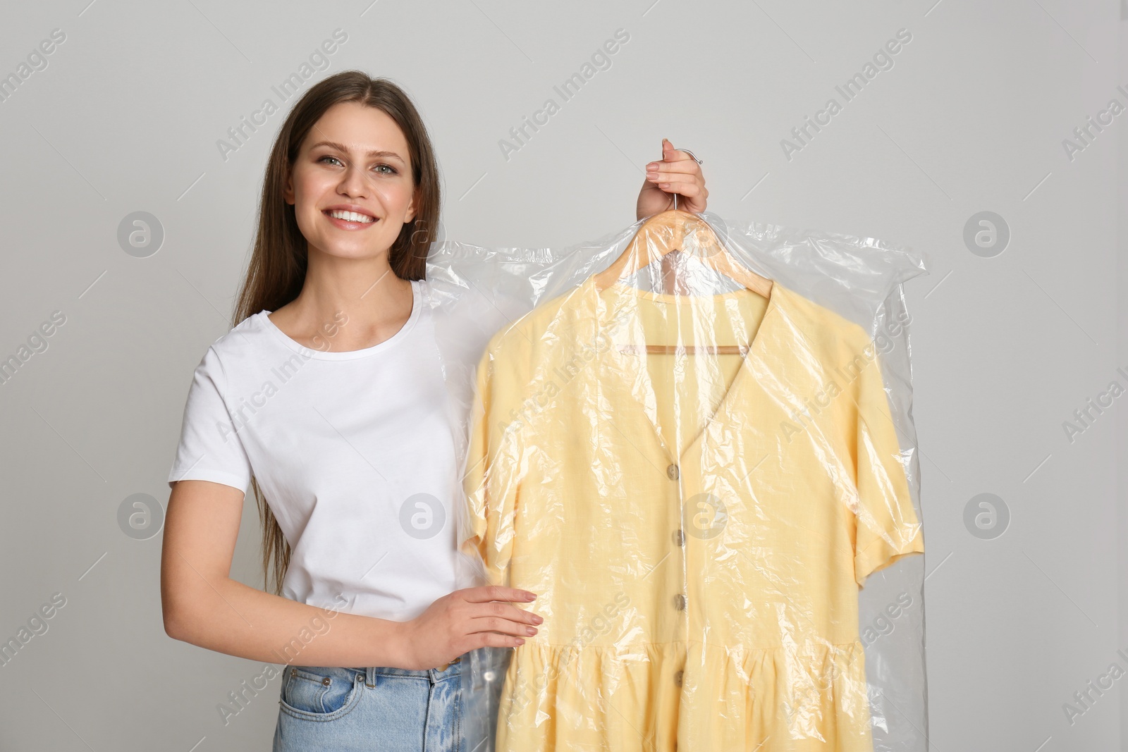 Photo of Young woman holding hanger with dress in plastic bag on light grey background. Dry-cleaning service