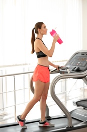 Photo of Athletic young woman drinking protein shake on running machine in gym