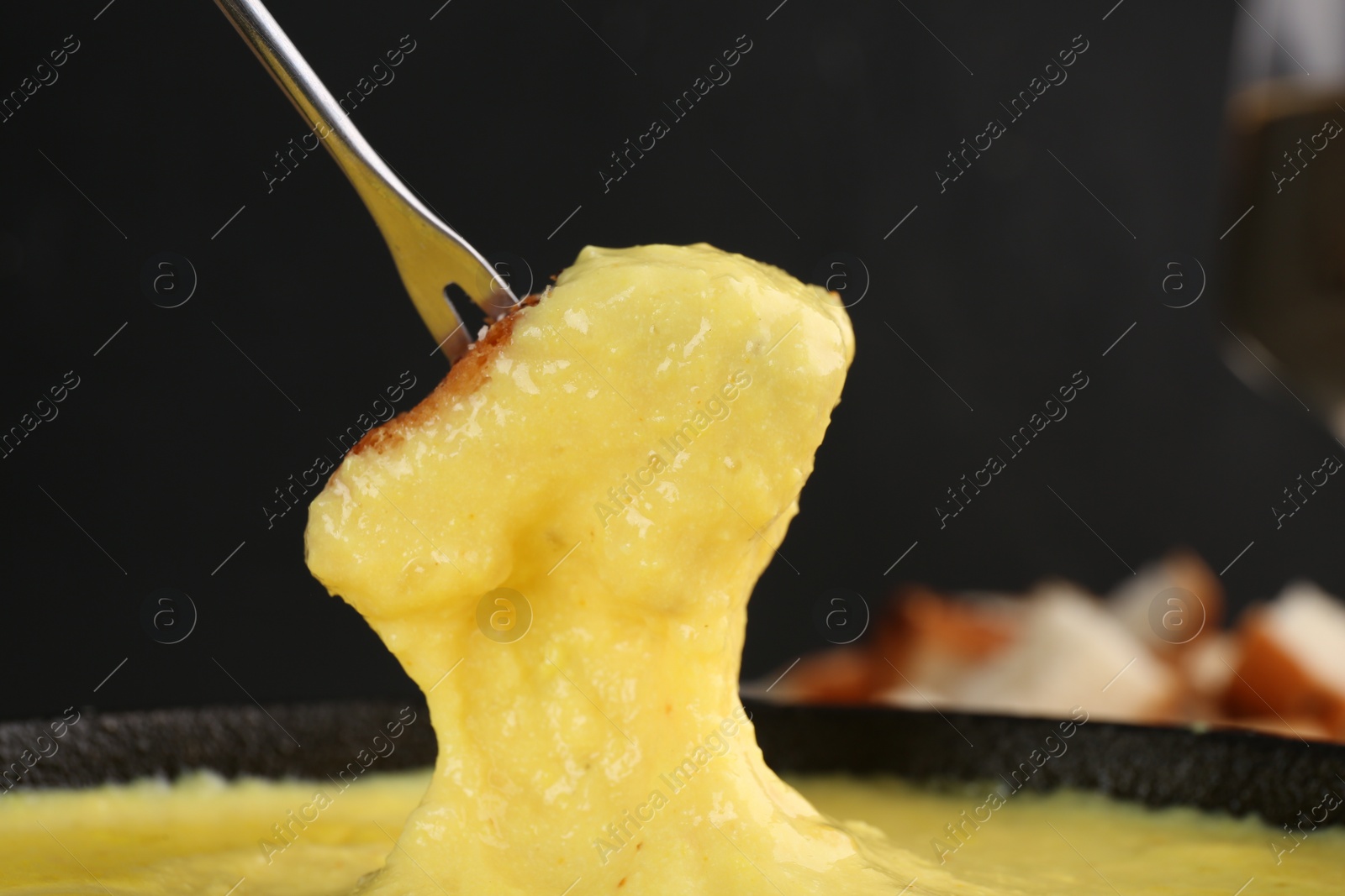 Photo of Dipping piece of bread into fondue pot with tasty melted cheese against dark gray background, closeup