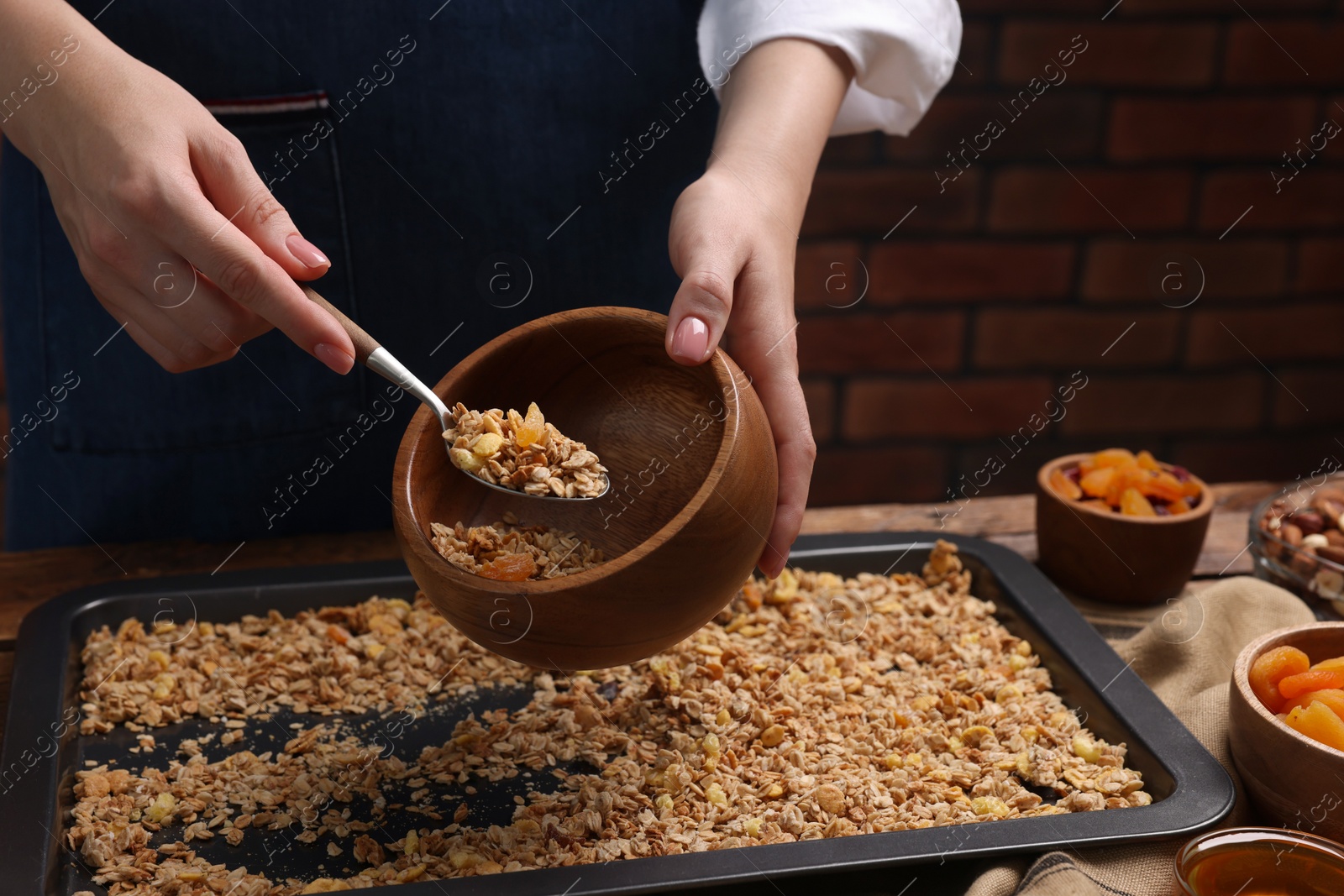 Photo of Woman putting granola from baking tray into bowl at table, closeup