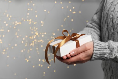 Christmas present. Woman holding gift box against grey background with blurred lights, closeup. Space for text