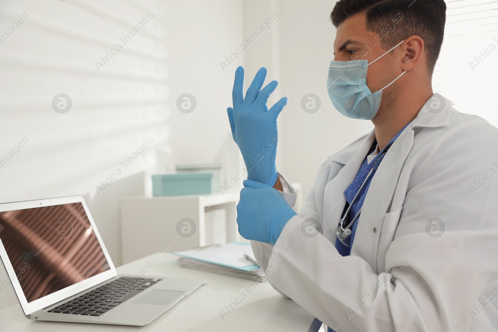 Photo of Doctor in protective mask putting on medical gloves at table in office