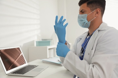 Doctor in protective mask putting on medical gloves at table in office