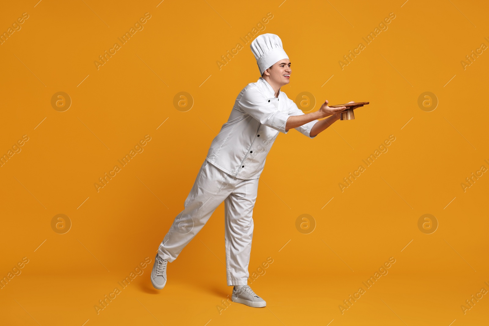 Photo of Portrait of happy confectioner in uniform holding cake stand on orange background