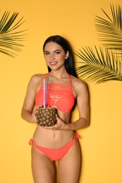 Beautiful young woman in stylish bikini with cocktail and tropical leaves on yellow background