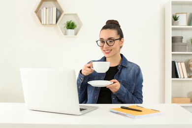 Home workplace. Happy woman with cup of hot drink looking at laptop at white desk in room