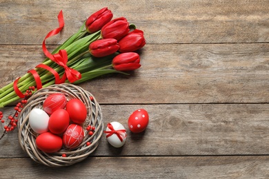 Photo of Flat lay composition of nest with painted Easter eggs and flowers on wooden table, space for text