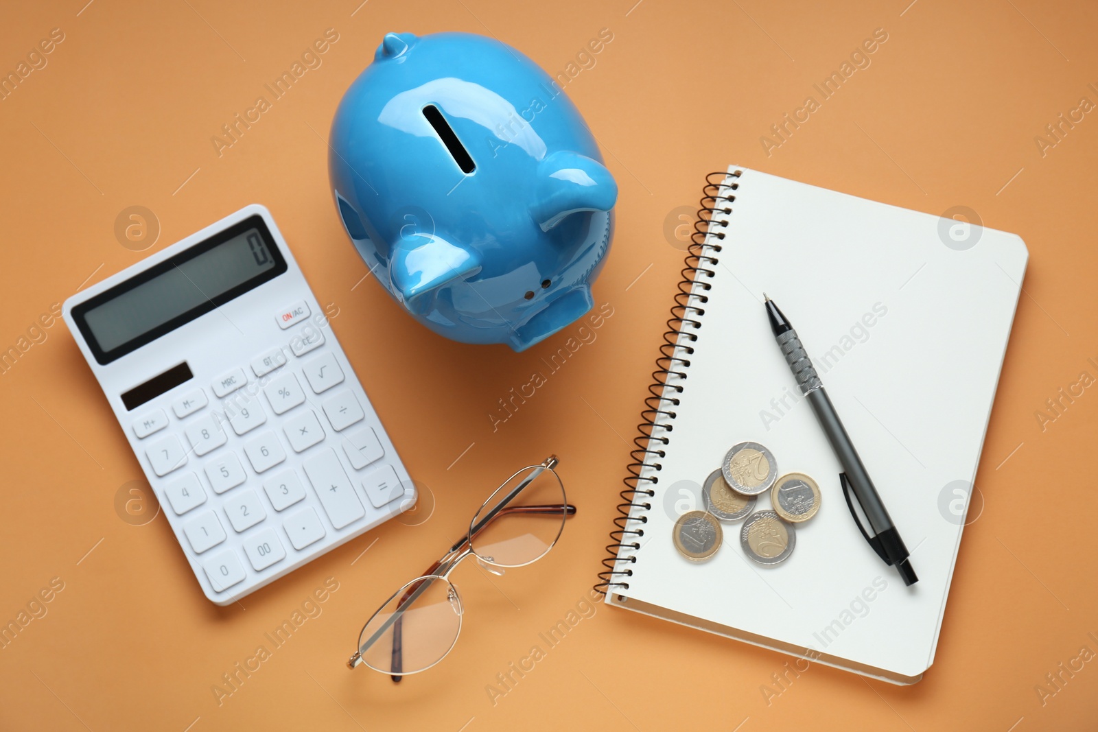 Photo of Piggy bank, notebook with coins, glasses and calculator on orange background, flat lay