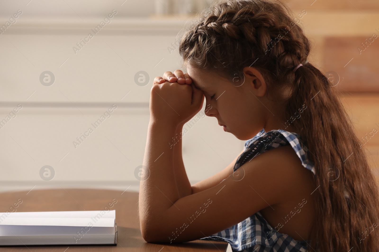 Photo of Cute little girl praying over Bible at table in room