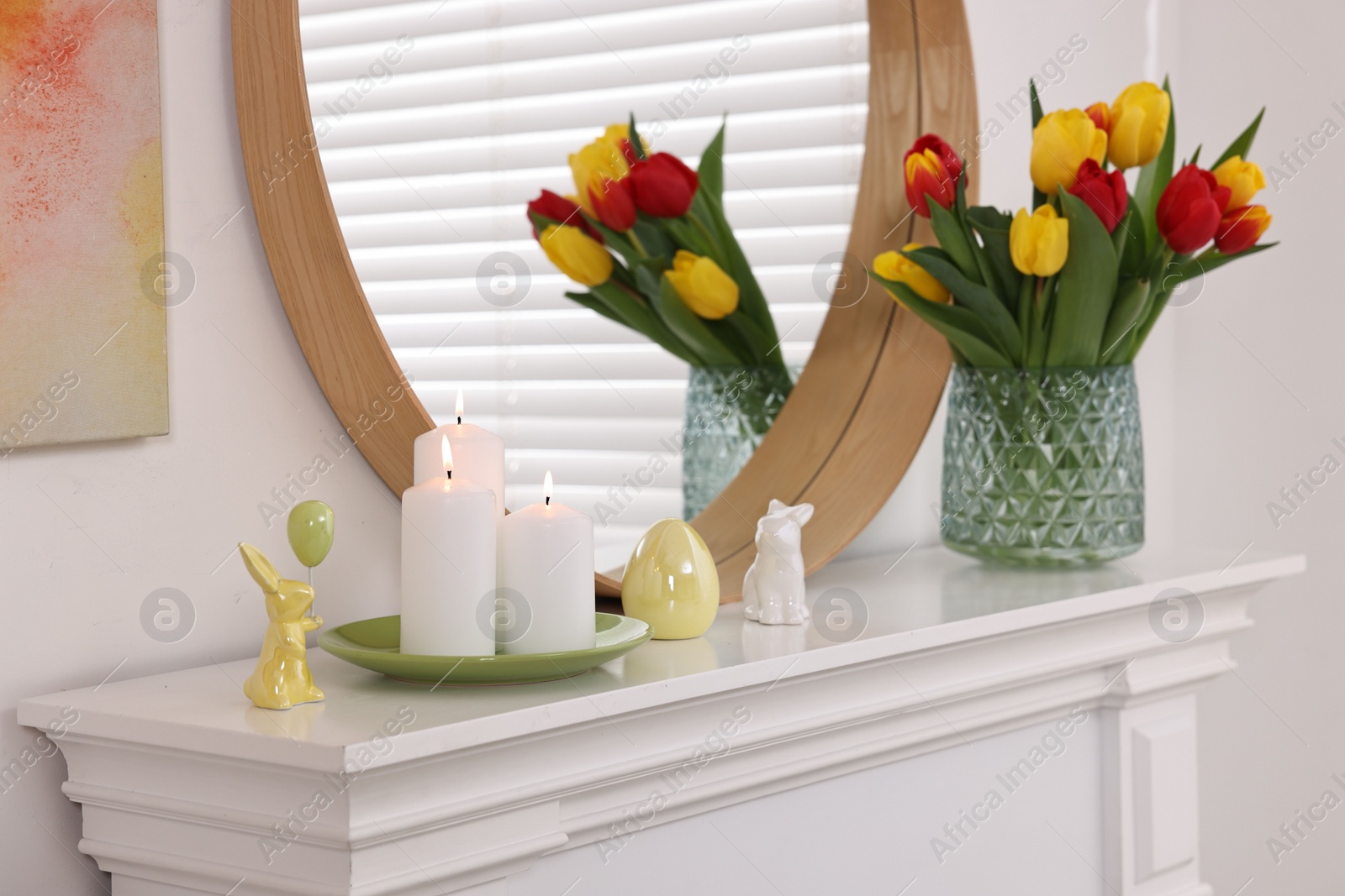 Photo of Easter decorations. Bouquet of tulips in vase, burning candles and bunny figures on fireplace at home