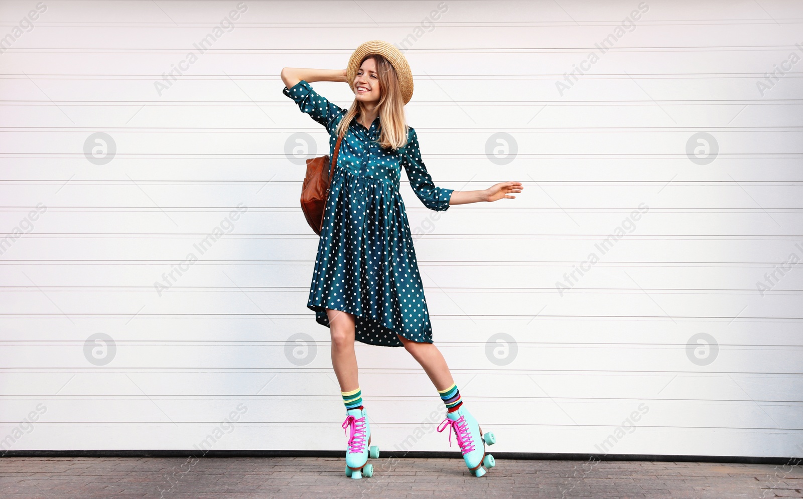 Photo of Happy stylish young woman with vintage roller skates, hat and backpack near white garage door on street