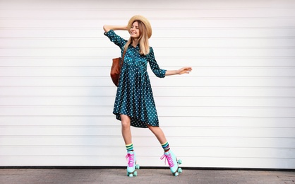 Happy stylish young woman with vintage roller skates, hat and backpack near white garage door on street