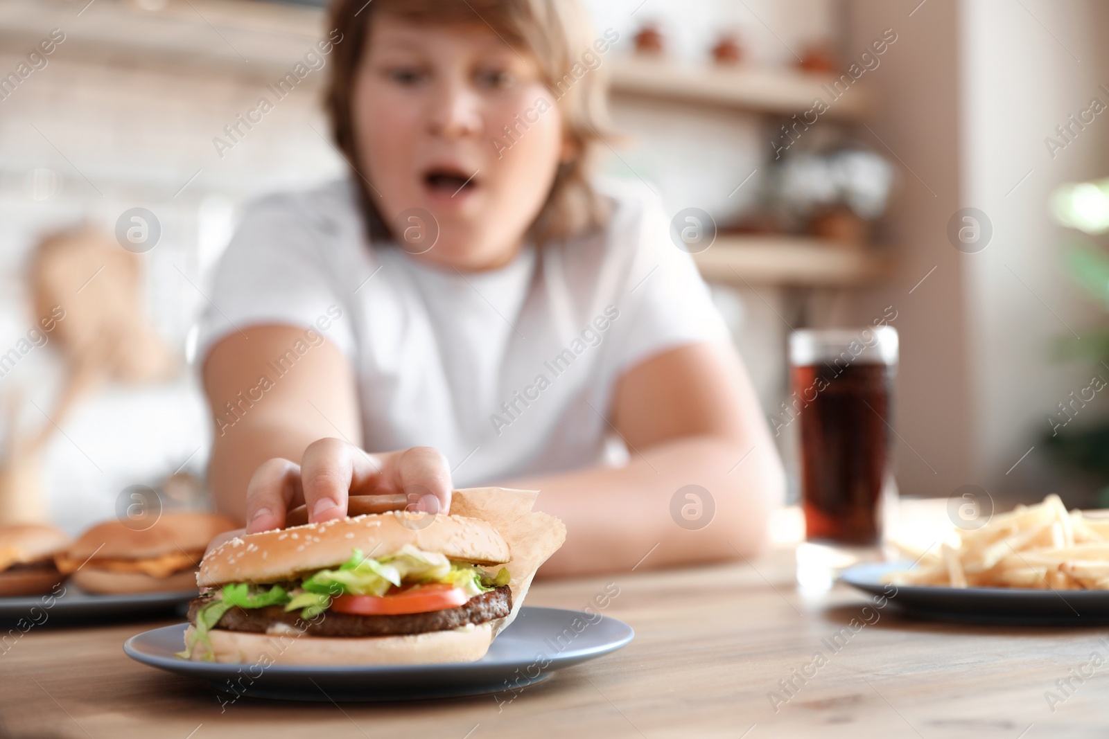 Photo of Emotional overweight boy reaching for burger at table in kitchen