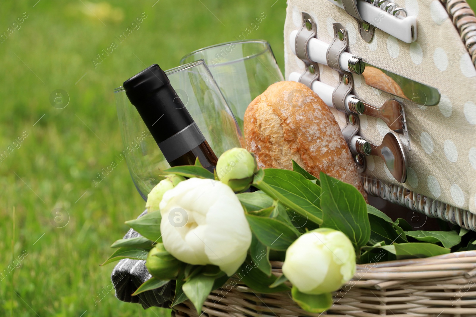 Photo of Picnic basket with wine, bread and flowers on green grass outdoors, closeup