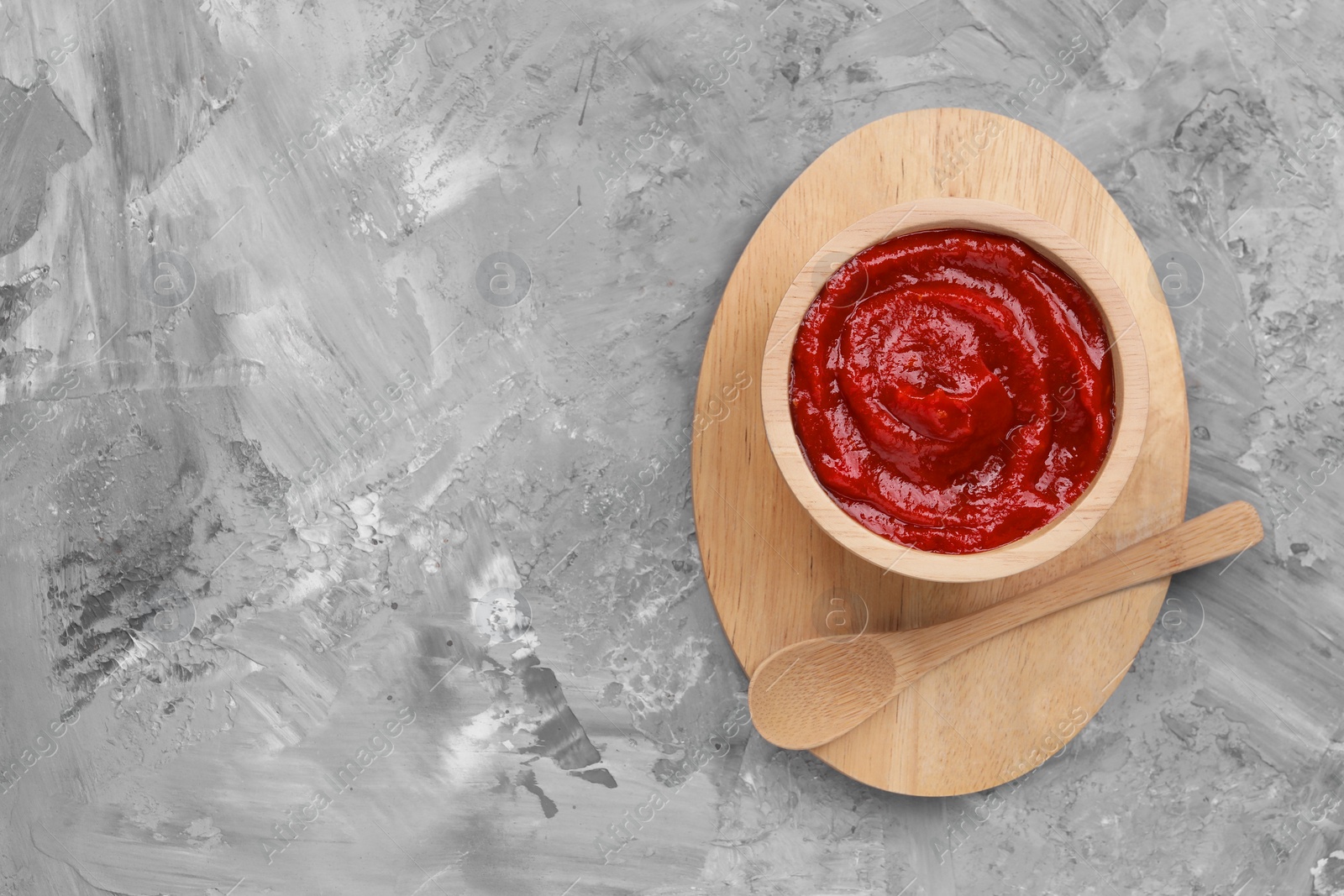 Photo of Organic ketchup in wooden bowl and spoon on grey textured table, top view with space for text. Tomato sauce